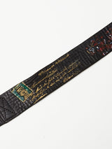 A close-up of the Gari Belt - Black by Blue de Gênes shows its black leather strap, gold text, vibrant embroidery with stitched detailing, a small green patch on the left, and floral designs on the right against a plain white background.