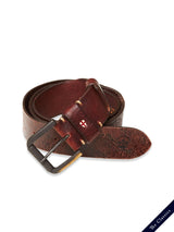 The Piceno Leather Belt in cognac by Blue de Gênes features subtle stitching, a silver buckle, and a red cross emblem. Its vintage look is highlighted against a plain white background with The Classics on a diagonal strip. Crafted from rolled-up brown cow leather.