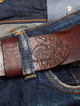 Close-up of blue jeans with orange stitching, showcasing the Blue de Gênes Piceno Leather Belt in cognac. The belt features a textured surface with an embossed design for a vintage look, and a logo-embossed label peeks at the top.