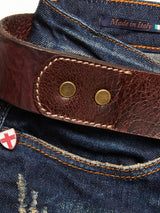 Close-up of Blue de Gênes blue denim jeans with orange stitching, paired with the Piceno Leather Belt in cognac. The belt has a logo-embossed buckle and a Made in Italy label. A small shield patch with a red cross near the pocket enhances the vintage look.