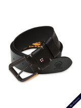 The Piceno Leather Belt - Black by Blue de Gênes, featuring a silver buckle and a small shield emblem with a cross, exudes a vintage charm. Displayed on a white background, it proudly bears The Classics label in the bottom corner.