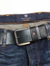 A close-up of dark blue denim jeans paired with the Piceno Leather Belt - Black by Blue de Gênes. The belt has a simple metallic buckle and subtle logo emblem. The jeans feature orange stitching, a vintage look, and a label with slightly blurred text on the waistband.