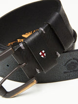 The Piceno Leather Belt - Black by Blue de Gênes showcases a close-up of black cow leather with a vintage texture, featuring a bronze buckle, stitched details, an embossed logo, and a small red and white cross emblem on the loop.