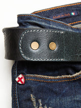 Close-up of Blue de Gênes blue denim jeans with orange stitching, featuring a Piceno Leather Belt in black with brass rivets and an embossed logo. The jeans have a vintage look accented by a small red and white shield pin, while inside, the label reads Made in Italy alongside an Italian flag.