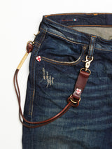 A pair of blue denim jeans with visible distressing and a BGsavino Key Hanger - Brown by Blue de Gênes, featuring metal clasps on the belt loop and accented with a small red and white shield emblem and logo rivet detail.
