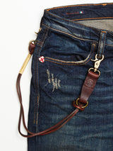 Close-up of Blue de Gênes blue denim jeans with distressed details, a logo rivet, red and white cross pin, and the BGsavino Key Hanger - Brown featuring a metal clasp on the belt loop. The jeans are Made in Italy.