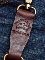 A close-up of the BGSavino Key Hanger in brown cow leather by Blue de Gênes, featuring embossed branding and a logo rivet, displayed on blue denim fabric.