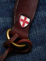 A close-up of the BGsavino Key Hanger - Brown by Blue de Gênes, crafted from cow leather with a gold ring and shield emblem featuring a red cross on white, rests on blue denim, highlighting the logo rivet detail.