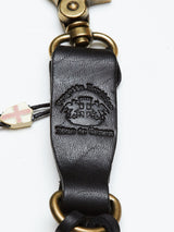 Close-up of the BGsavino Key Hanger in black cow leather by Blue de Gênes, featuring embossed branding, brass hardware, and a small charm with a red cross on white. The strap displays "Prosotto Tecnico" and "Blue de Gênes".