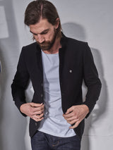 A man with long hair and a beard adjusts a button on his Blue de Gênes Casella Officers Blazer - Black over a light blue shirt, set against a light-colored wall. Paired with jeans, the brushed cotton fabric adds textured charm for a casual yet stylish look.