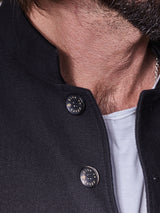 A person is wearing a Casella Officers Blazer - Black by Blue de Gênes. The blazers vintage parade uniform style with two Blue Rouge buttons is layered over a white t-shirt. The brushed cotton adds texture, and a glimpse of silver necklace chain and facial hair are visible.
