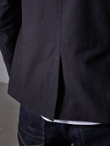 A person in dark blue jeans and a Black Casella Officers Blazer by Blue de Gênes is shown. The focus is on the blazers back slit, made from brushed cotton. Visible jean stitching and a glimpse of a white shirt are under the blazer against a neutral backdrop.