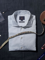 A neatly folded white button-up shirt, crafted from Italian cotton fabric, rests on a dark surface. A measuring tape is draped over the Miguel Brilliante Shirt by Blue de Gênes. Nearby, a Corozo nut button and a label reading The Classics add detail.