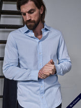 A man with a beard stands indoors, looking downward thoughtfully while adjusting his cuffs. He wears the Miguel Brilliante Shirt in light blue, crafted from fine Italian cotton by Blue de Gênes. A ladder is visible in the background with The Classics text in the corner.