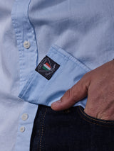 A person is wearing a Miguel Brilliante Shirt - Light Blue by Blue de Gênes, made from fine Italian fabric. The light blue cotton shirt has white buttons, and part of the visible black label says Italy. The person’s hand is partially tucked into the pocket of dark jeans.