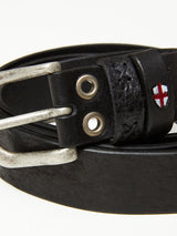 Close-up of the Barra Belt - Black by Blue de Gênes, featuring black cow leather with a silver buckle, two metal eyelets, vintage look, textured finish with exquisite handmade stitch details, and a small emblem displaying a red cross on a white background.