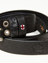 Close-up of the Barra Belt - Black by Blue de Gênes, crafted from black cow leather with embossed details, eyelets, and a small patch featuring a red cross on white. Handmade stitching and a decorative emblem provide a subtle vintage appeal.