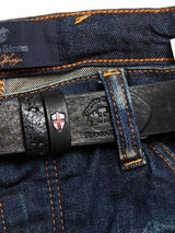 Close-up of dark blue denim jeans with orange stitching, paired with the Blue de Gênes Barra Belt - Black made of cow leather; it features an embossed logo and a red and white shield symbol, adding a vintage touch.