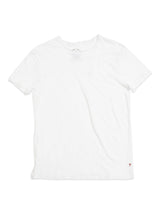 The Lino T-shirt by Blue de Gênes is a plain white T-shirt made from lightweight linen with short sleeves and a round neckline. It features a small red cross logo near the bottom right hem and unique raw edge details for added style.