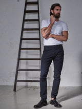 A tattooed man stands thoughtfully near a tall wooden ladder against a plain wall, wearing a garment-dyed white t-shirt and Blue de Gênes Teo Chino in navy.