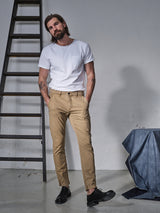 A man with long hair and a beard stands casually, his hands in the pockets of his Teo Chino - Antique Bronze pants by Blue de Gênes. He wears a plain white t-shirt and black shoes. Behind him, a ladder and draped blue cloth complete the vintage look.