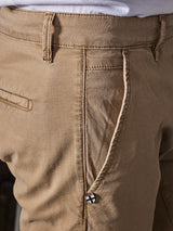 Close-up of someone wearing Blue de Gênes Teo Chino in Antique Bronze, featuring a visible zipper and a small cross charm. The textured honeycomb fabric has a vintage appearance, with a partly visible side pocket.