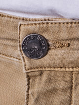 Close-up of the Teo Chino in Antique Bronze by Blue de Gênes. The metal button, embossed with decorative designs and Blue de Gênes, enhances the vintage look. Light brown stitching is visible, and a white garment peeks above the waistband.