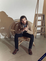 A man with long hair and a beard sits in a round wicker chair, wearing a garment dyed tan jacket over a white shirt and Blue de Gênes Teo Chinos in navy. Hes holding a phone, with a ladder and an easel behind him, as playful shadows dance on the wall.