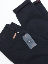 Two pairs of navy Teo Chino trousers by Blue de Gênes are folded on a light surface. One pair has a label with a button showing branding details. The garment-dyed trousers feature side pockets and belt loops.