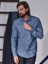 A bearded man with long hair, wearing the Blue de Gênes Miguel Chambray Shirt in Lt. Denim Blue and dark jeans, stands near a ladder with folded clothes, looking down with his hand near the shirt collar. The text The Classics is in the bottom corner.