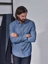 A man with long hair and a beard stands with arms crossed, wearing the Blue de Gênes Miguel Chambray Shirt in Lt. Denim Blue paired with jeans. The elegant corozo nut buttons enhance his look, while a denim jacket drapes over a ladder in the background for a relaxed yet stylish vibe.