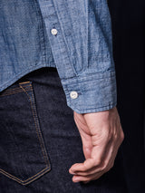 A person is wearing a Miguel Chambray Shirt - Lt. Denim Blue by Blue de Gênes, featuring Corozo nut buttons, paired with dark blue jeans showcasing orange stitching on the back pocket.