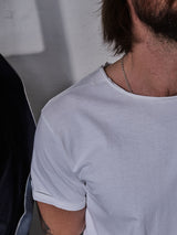 A person with shoulder-length brown hair, wearing a Blue de Gênes Sagi Nuovo T-shirt in white and a silver chain necklace, is partially visible. The premium cotton shirt complements their style as they sit against a light wall with shadows around them, sporting a dark jacket draped over one shoulder.