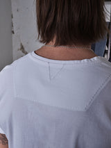 The image shows someone with shoulder-length brown hair wearing a regular fit Blue de Gênes Sagi Nuovo T-shirt in white, crafted from premium cotton with refined stitching, accompanied by a silver chain necklace. A wall with peeling paint appears in the background.