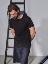 A man with long hair and a beard leans against a wooden ladder, wearing the Blue de Gênes Sagi Nuovo T-shirt - Black made of premium cotton and regular fit jeans. The simple setting features a white wall labeled The Classics, capturing timeless wardrobe essentials.