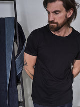 A man with long hair and a beard stands pensively in front of a ladder, wearing the Sagi Nuovo T-shirt - Black by Blue de Gênes. A blue denim garment, a true wardrobe staple, hangs over the ladder as he gazes off to the side.
