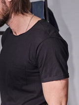 A person with shoulder-length hair and a beard wears the Sagi Nuovo T-shirt by Blue de Gênes—a wardrobe staple with a left-side pocket. Made from premium cotton, this regular-fit black tee reveals a chain necklace as they face slightly away from the camera.