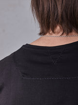 The image displays a person from behind wearing a Blue de Gênes Sagi Nuovo T-shirt in black, crafted from premium cotton with triangular stitching. A silver chain necklace accents the look alongside shoulder-length brown hair, all set against a gray background.