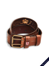 The Cintura Belt - Cognac by Blue de Gênes is a vintage-style brown cow leather military belt with a striking gold spiral double buckle, featuring a regal crest inside and a red-and-white cross patch on the loop. It has a navy tag labeled The Classics at the bottom right corner.