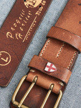 Close-up of the Cintura Belt - Cognac by Blue de Gênes on a striped denim surface. This Swiss-flagged, engraved, vintage military belt features a metallic double buckle and comes in size L.