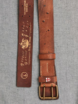 The Cintura Belt - Cognac by Blue de Gênes is crafted from brown cow leather. It features a double buckle with engraved sailing ship designs, Poetic Queen and Made in Italy. A small emblem with a red cross on a white background near the brass buckle adds a vintage military touch.