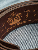 Close-up of the Cintura Belt - Cognac by Blue de Gênes, on a textured blue fabric. The vintage-style brown leather belt has a worn logo, decorative golden markings, and an illustrated ship design.