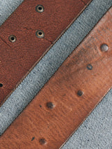 Close-up of two Cintura Belts in cognac by Blue de Gênes on a textured gray fabric. The top belt, with circular eyelets, resembles a vintage military style, while the bottom belt displays wear with visible indentations.