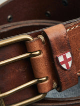 The Cintura Belt - Cognac by Blue de Gênes is a vintage military-style belt, crafted from brown cow leather and featuring a brass double buckle. It displays a small emblem of a red cross on a white background, with visible stitching that enhances its durability and style.