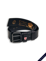 The Cintura Belt - Black by Blue de Gênes is a vintage leather belt with a silver buckle featuring a red and white shield emblem. Rolled, it reveals embossed gold lettering inside. A small tag reads The Classics in white on dark blue, adding to its timeless appeal.