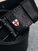 A close-up of the Cintura Belt - Black by Blue de Gênes, featuring white stitching and a small shield emblem with a red cross, rests on a blue denim fabric background, highlighting its military-inspired design.