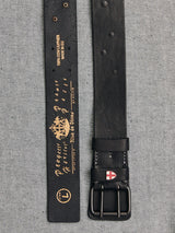 A close-up of the Cintura Belt - Black by Blue de Gênes on a gray textured surface shows two sections: one with intricate gold designs and text, and the other featuring a double buckle with a red and white emblem, inspired by military style.