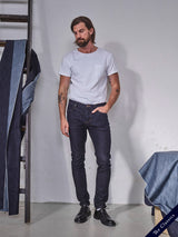 A man with long hair and a beard wears casual attire, including a white t-shirt, Repi Dai V Dark Jeans by Blue de Gênes, and black shoes. He poses beside hanging denim fabric and a table with folded cloth from Repi Jeans against a plain studio wall.