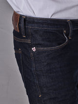 A close-up shows someone sporting Blue de Gênes Repi Dai V Dark Jeans in dark blue denim, featuring brown stitching and a brown leather patch on the waistband. A red cross pin accents the front pocket of these regular slim-fit jeans, paired with a crisp white shirt.