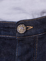 Close-up of Repi Dai V Dark Jeans by Blue de Gênes, featuring slim-fit dark blue denim with comfort stretch. Detailed yellow stitching and a metal button with ship designs complement the jeans, worn over a crisp white shirt.